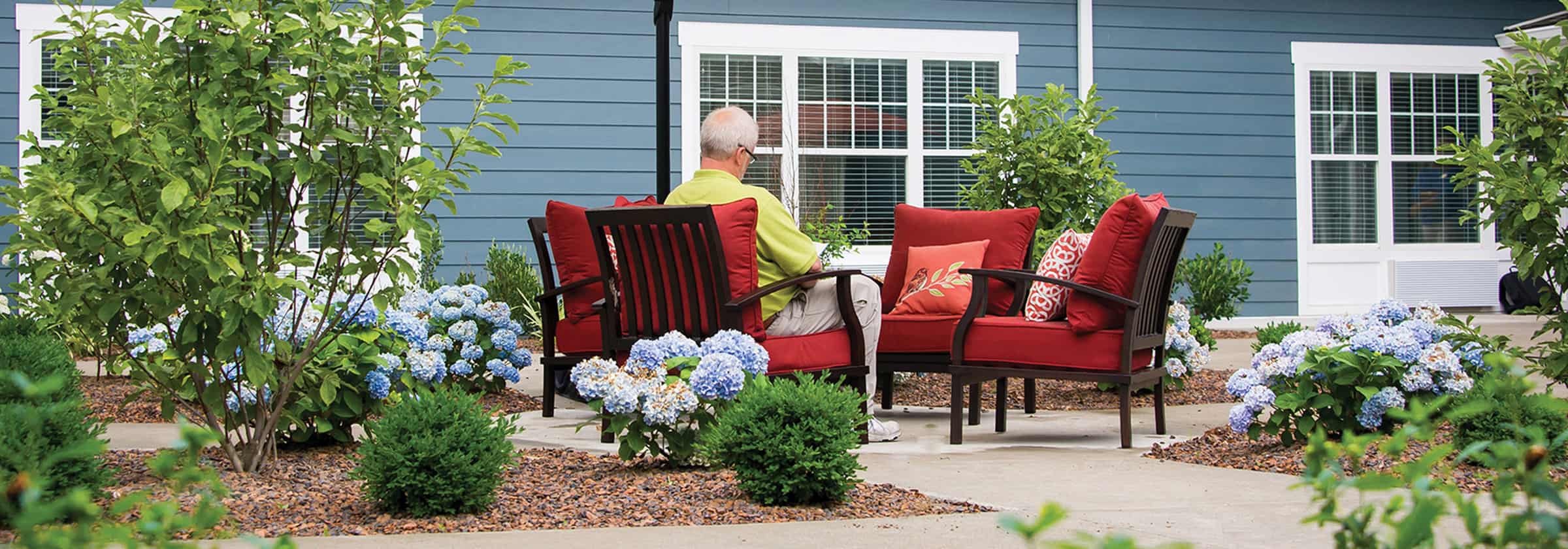 A senior man sitting in a black metal outdoor chair with red cushions reading on a patio in front of a blue senior living apartment building