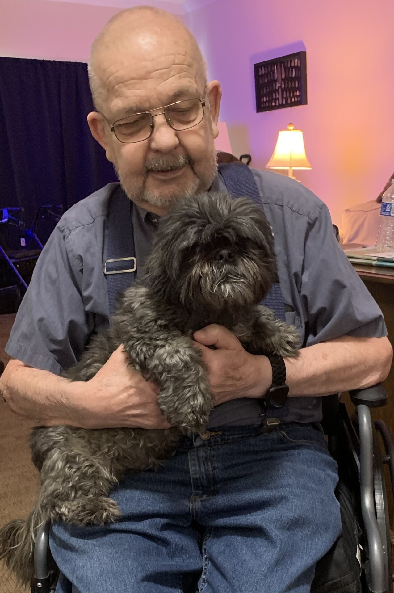 An elderly resident of a Traditions Management senior living facility holding a small long-haired black dog on his lap
